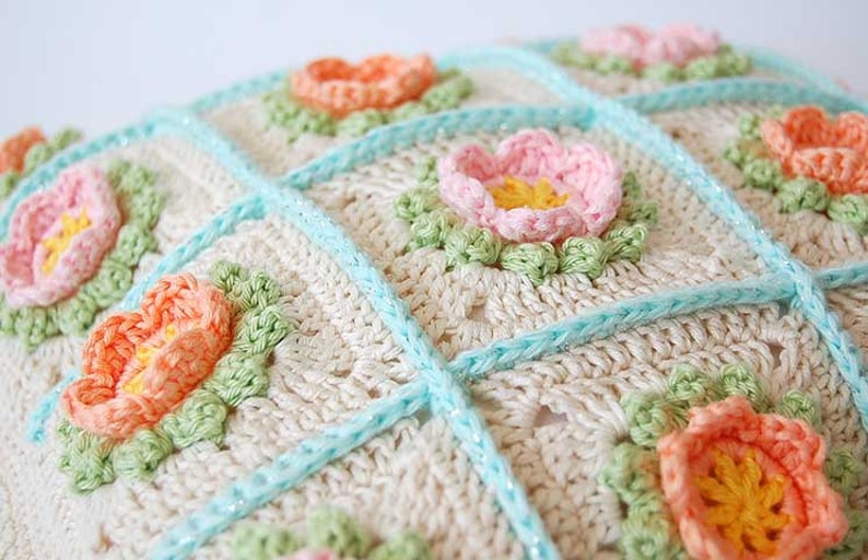 May include: A crocheted pillow with a white background and a pattern of pastel pink, peach, and yellow flowers. The pillow is trimmed with a light blue border.