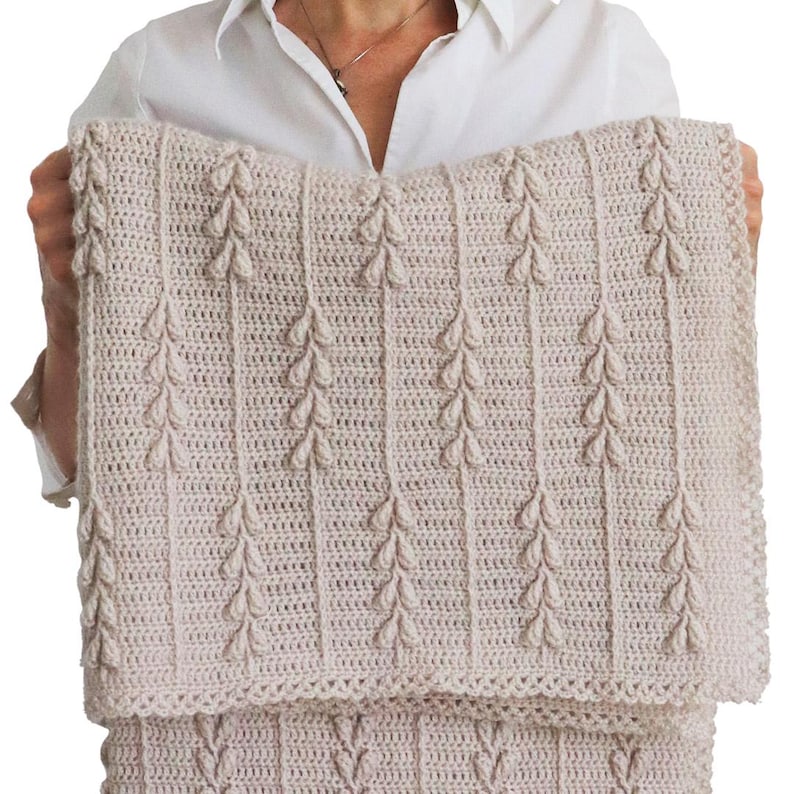 Puede incluir: Una manta rectangular de ganchillo en color beige claro con un patr&oacute;n vertical repetitivo de racimos en relieve con forma de hoja. La textura est&aacute; creada con puntos densos y los bordes est&aacute;n rematados con un delicado borde de ganchillo festoneado.