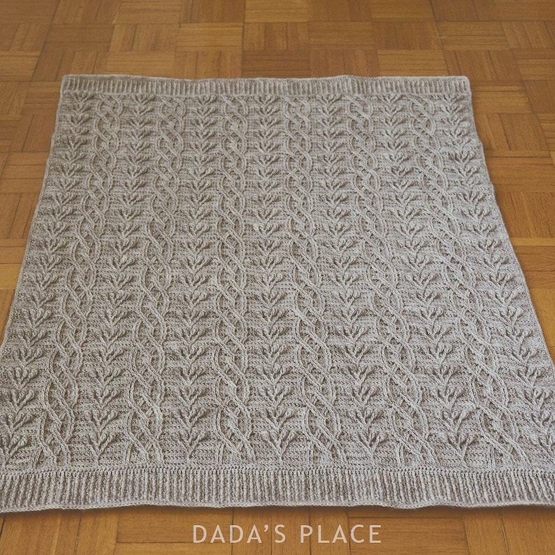 May include: A beige knitted blanket with a repeating leaf pattern. The blanket has a ribbed border and is displayed on a wooden floor. The text "DADA'S PLACE" is visible in the bottom right corner.