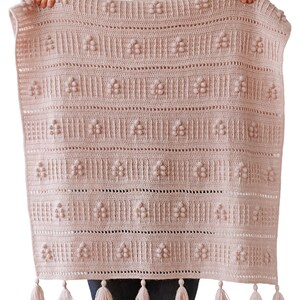 Peut inclure: Une couverture en crochet rose avec un motif floral et des franges. La couverture est tenue par les mains d'une personne. Le texte "DADA'S PLACE" est visible dans le coin inf&eacute;rieur droit de l'image.