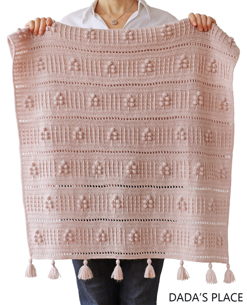 Peut inclure: Une couverture en crochet rose avec un motif floral et des franges. La couverture est tenue par les mains d'une personne. Le texte "DADA'S PLACE" est visible dans le coin inf&eacute;rieur droit de l'image.