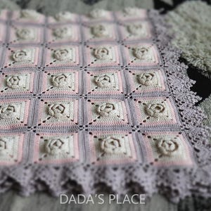 Op de afbeelding: Een handgemaakte gehaakte deken met een herhalend patroon van vierkante blokken met verhoogde rozenontwerpen. De deken is in roze, crème en grijstinten, met een geschulpte rand. De tekst "DADA'S PLACE" is zichtbaar aan de onderkant.