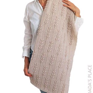 Puede incluir: Una manta rectangular de ganchillo en color beige con un patr&oacute;n vertical repetitivo de puntos en relieve en forma de hoja, separados por columnas de puntos altos. El tejido tiene un color tostado uniforme e incluye el texto DADA'S PLACE.