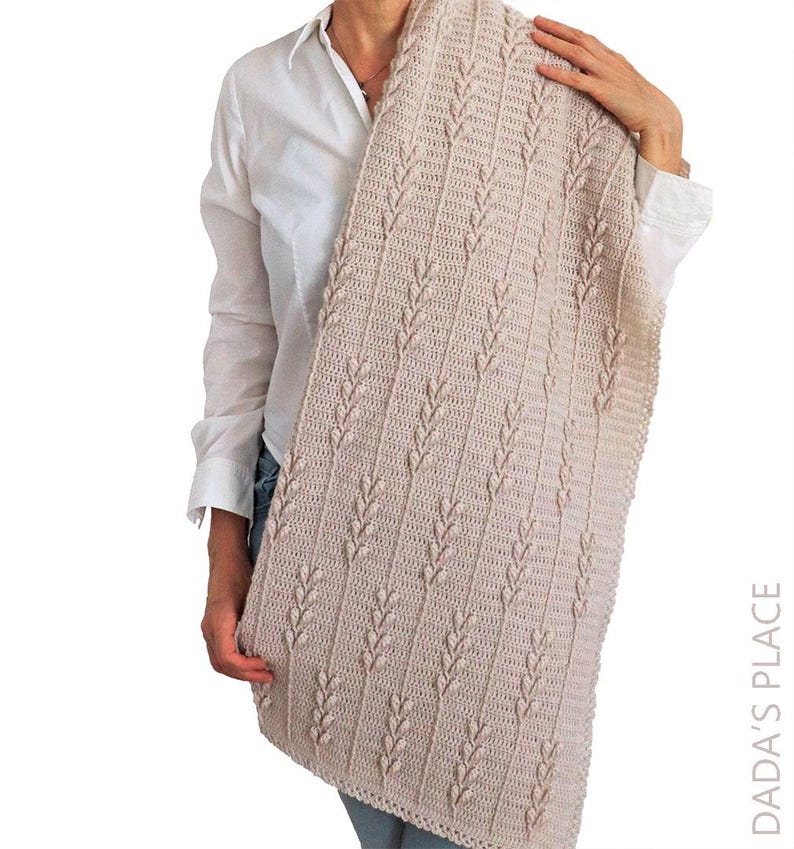 Puede incluir: Una manta rectangular de ganchillo en color beige con un patr&oacute;n vertical repetitivo de puntos en relieve en forma de hoja, separados por columnas de puntos altos. El tejido tiene un color tostado uniforme e incluye el texto DADA'S PLACE.