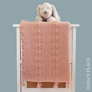 May include: A pink crocheted baby blanket with a textured leaf pattern and tassels. The blanket is draped over a white dresser with a stuffed animal on top.