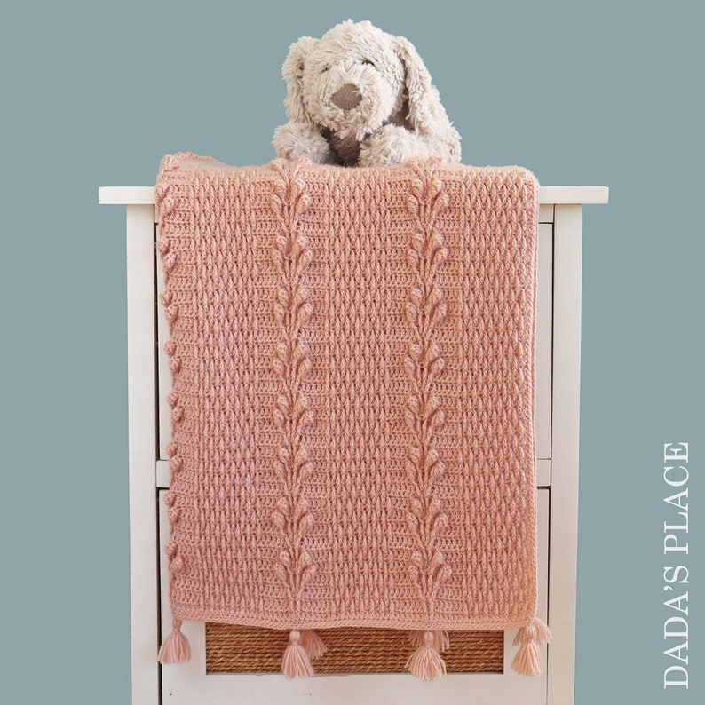 May include: A pink crocheted baby blanket with a textured leaf pattern and tassels. The blanket is draped over a white dresser with a stuffed animal on top.