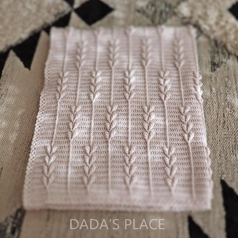Puede incluir: Una manta rectangular de ganchillo de color rosa p&aacute;lido con un patr&oacute;n vertical repetitivo de tallos elevados similares al trigo. Los bordes est&aacute;n rematados con un delicado borde festoneado. El texto DADA'S PLACE est&aacute; impreso en blanco.