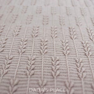 Puede incluir: Una manta de ganchillo de color beige claro con un patr&oacute;n vertical repetitivo de tallos texturizados en relieve que recuerdan al trigo. El tejido tiene una construcci&oacute;n de puntos densa y uniforme. El texto DADA'S PLACE est&aacute; impreso en blanco en la parte inferior central.