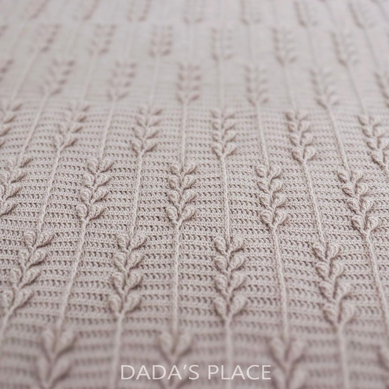 Puede incluir: Una manta de ganchillo de color beige claro con un patr&oacute;n vertical repetitivo de tallos texturizados en relieve que recuerdan al trigo. El tejido tiene una construcci&oacute;n de puntos densa y uniforme. El texto DADA'S PLACE est&aacute; impreso en blanco en la parte inferior central.