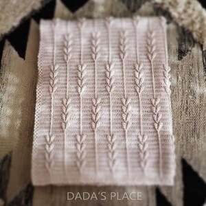 Puede incluir: Una manta rectangular de ganchillo en color rosa claro con un patr&oacute;n vertical repetitivo de tallos texturizados en relieve que recuerdan al trigo. El tejido tiene una base de punto fino y uniforme con un borde delicado. El texto DADA'S PLACE aparece abajo.