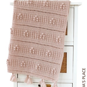 Peut inclure: Une couverture pour b&eacute;b&eacute; en crochet rose avec un motif textur&eacute; et des pompons. La couverture est pos&eacute;e sur une commode blanche.