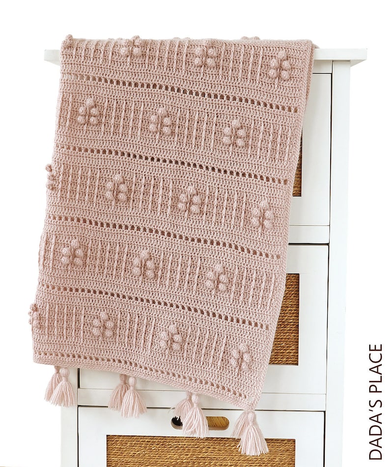Peut inclure: Une couverture pour b&eacute;b&eacute; en crochet rose avec un motif textur&eacute; et des pompons. La couverture est pos&eacute;e sur une commode blanche.