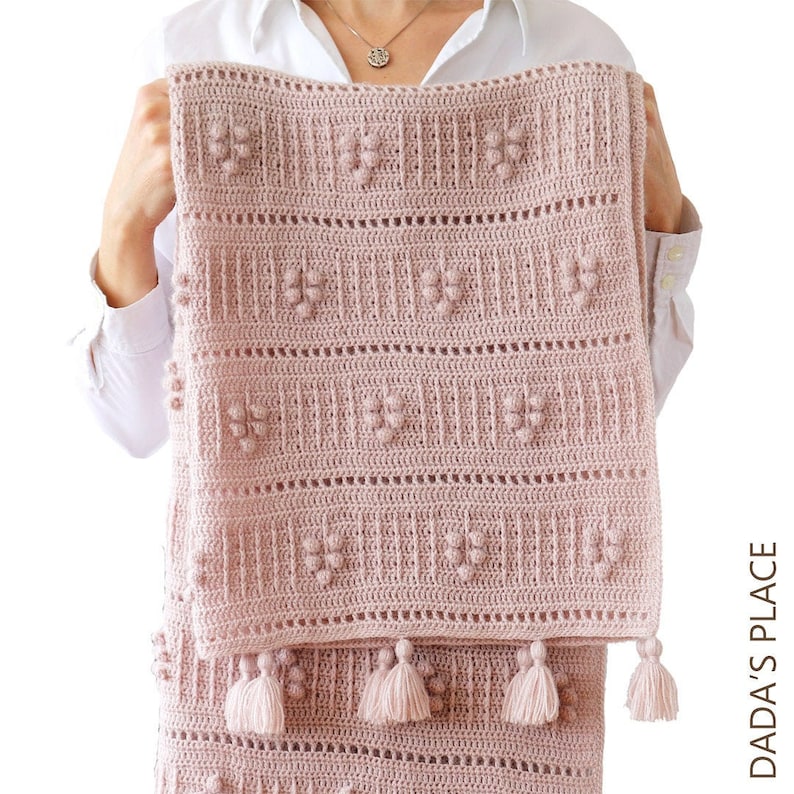 Peut inclure: Une couverture pour b&eacute;b&eacute; en crochet rose avec un motif textur&eacute; et des pompons sur les bords. La couverture est tenue par une personne portant une chemise blanche.