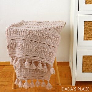 Peut inclure: Une couverture en crochet de couleur beige clair avec un motif textur&eacute; et des franges. La couverture est pos&eacute;e sur un tabouret en bois.
