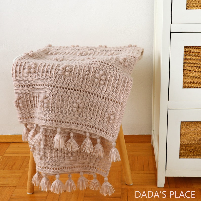 Peut inclure: Une couverture en crochet de couleur beige clair avec un motif textur&eacute; et des franges. La couverture est pos&eacute;e sur un tabouret en bois.