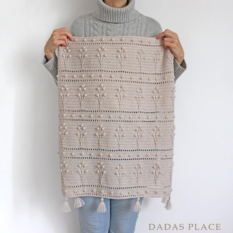 May include: A beige crochet blanket with a floral pattern and tassels. The blanket is held by a person wearing a grey turtleneck sweater and blue jeans. The text "DADAS PLACE" is visible in the bottom right corner of the image.