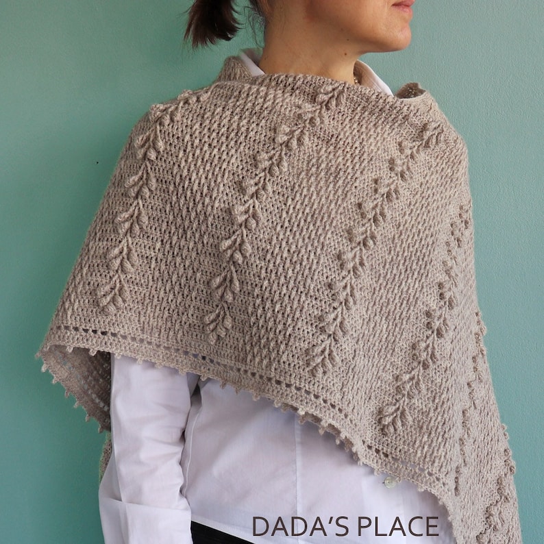 May include: A beige crochet shawl with a delicate leaf pattern and a scalloped edge. The shawl is draped over a person's shoulders. The text "DADA'S PLACE" is visible at the bottom of the image.