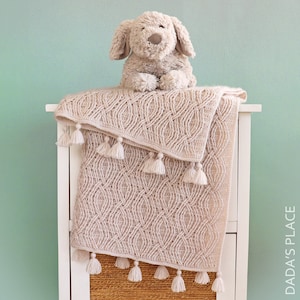 May include: A beige crocheted blanket with a repeating oval pattern and white tassels along the edges. A plush dog toy sits on top of the blanket. The blanket is draped over a white cabinet.