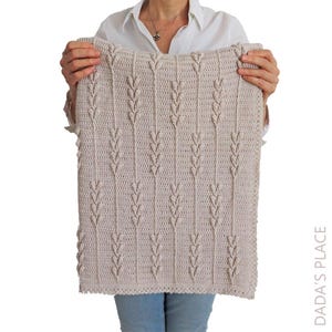 Puede incluir: Una manta rectangular de ganchillo en color beige claro con un patr&oacute;n vertical repetitivo de puntos texturizados en relieve que recuerdan a espigas de trigo. El tejido tiene una apariencia de punto uniforme y el texto DADA'S PLACE es visible.