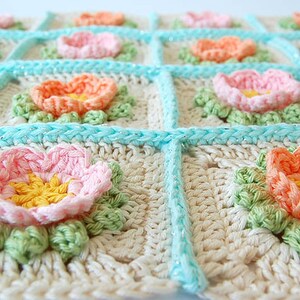 May include: A crocheted blanket with a floral design. The blanket is made of white yarn with light blue, pink, and orange flowers. The flowers are surrounded by green leaves.