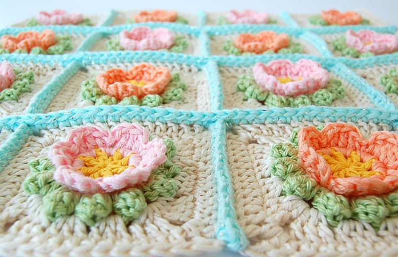 May include: A crocheted blanket with a floral design. The blanket is made of white yarn with light blue, pink, and orange flowers. The flowers are surrounded by green leaves.