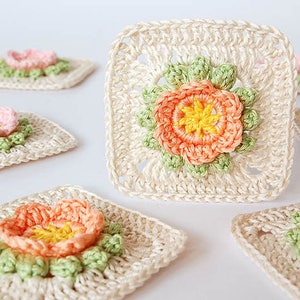 May include: Seven crocheted squares with a flower design in the center. The flowers are made with pink, peach, and yellow yarn. The squares are made with cream-colored yarn.