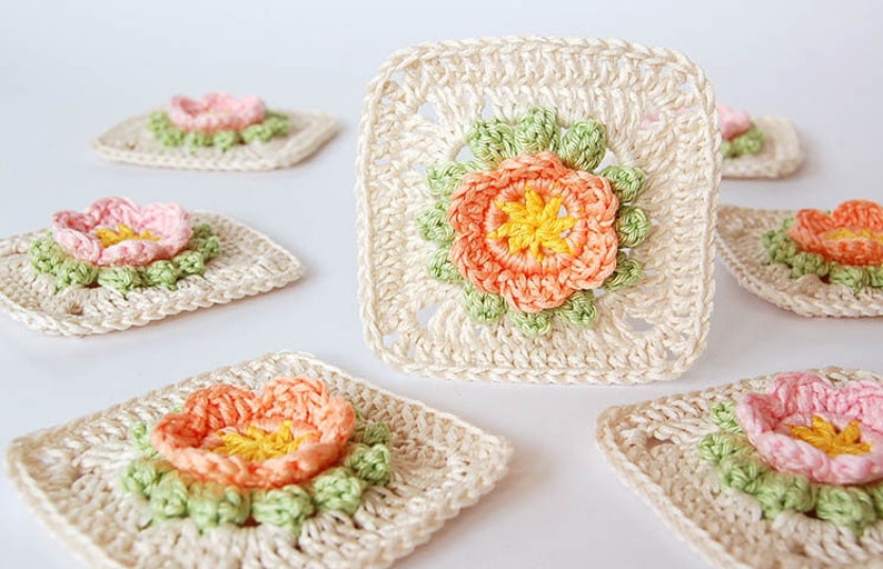 May include: Seven crocheted squares with a flower design in the center. The flowers are made with pink, peach, and yellow yarn. The squares are made with cream-colored yarn.