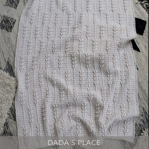 Puede incluir: Una manta de ganchillo color crema con un patr&oacute;n vertical repetitivo de puntos en relieve similares a hojas. El tejido tiene una superficie texturizada con un dise&ntilde;o de punto consistente. El texto DADA'S PLACE es visible en la parte inferior.