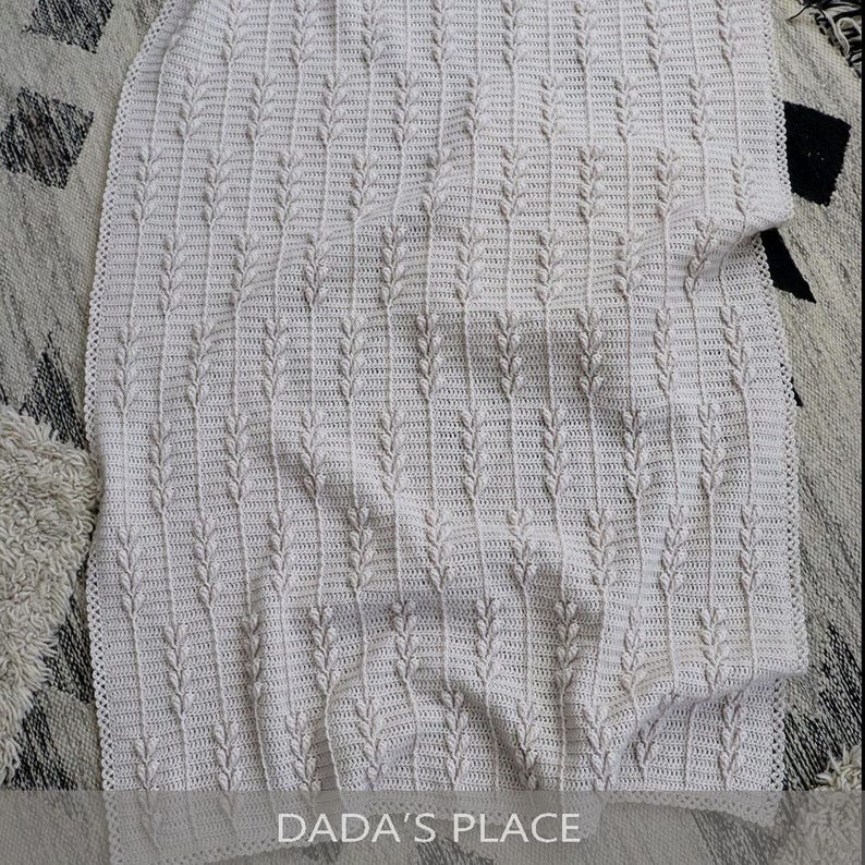 Puede incluir: Una manta de ganchillo color crema con un patr&oacute;n vertical repetitivo de puntos en relieve similares a hojas. El tejido tiene una superficie texturizada con un dise&ntilde;o de punto consistente. El texto DADA'S PLACE es visible en la parte inferior.