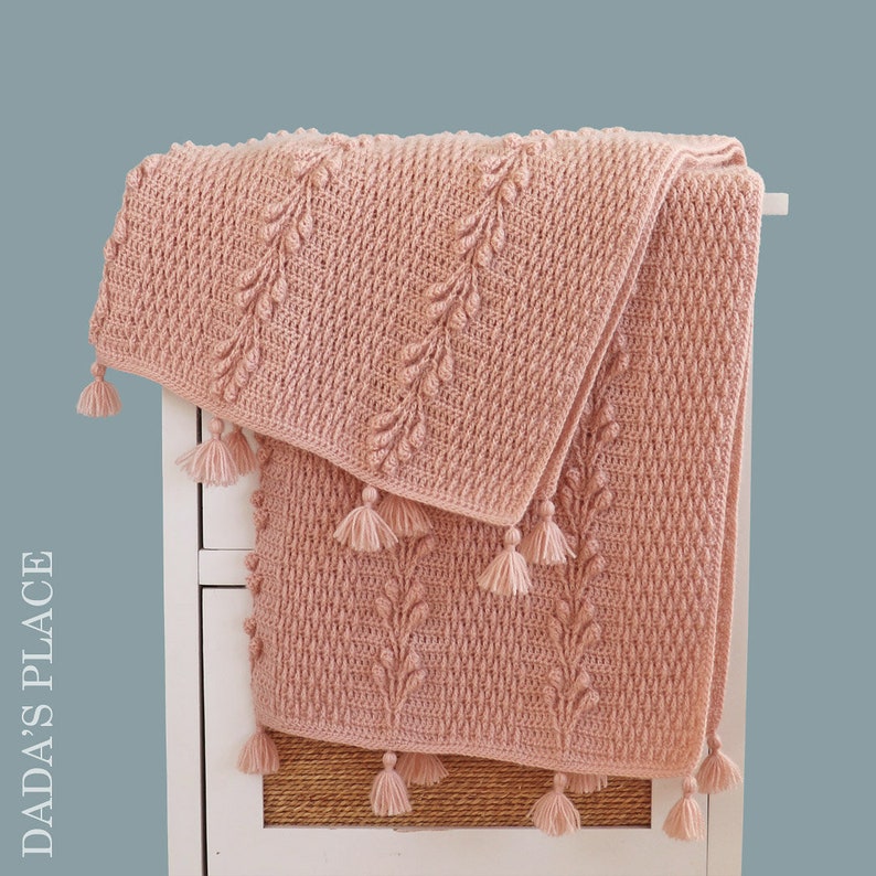 May include: A soft pink crocheted throw blanket with a textured leaf pattern and tassels. The blanket is folded and draped over a white shelf.