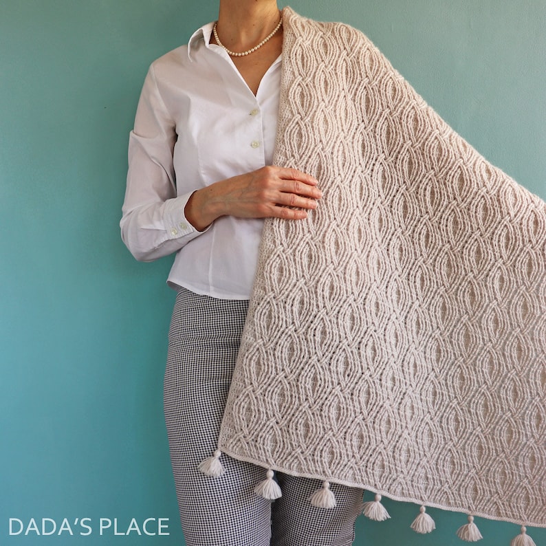 May include: A cream-coloured knitted shawl featuring a repeating oval design and white tassels. The shawl is draped over a person wearing a white shirt and black and white checked trousers. The background is a teal colour.