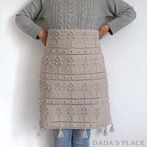 May include: A beige crochet skirt with a floral pattern and tassels. The skirt is made with a textured yarn and has a simple, elegant design.