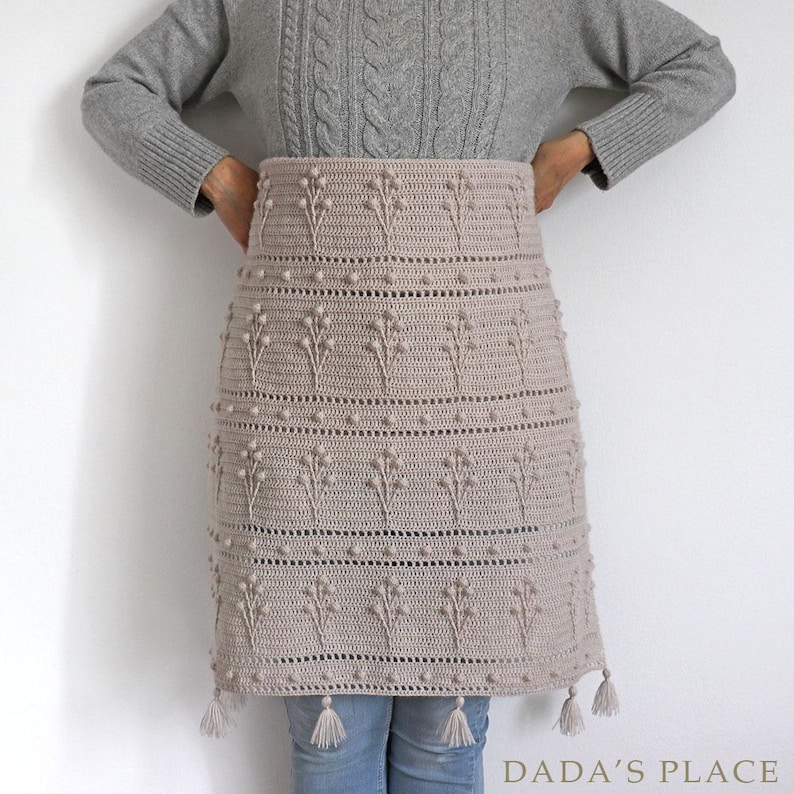 May include: A beige crochet skirt with a floral pattern and tassels. The skirt is made with a textured yarn and has a simple, elegant design.