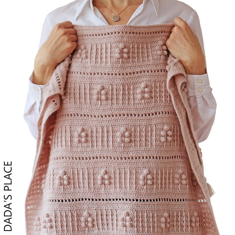 Peut inclure: Une couverture pour b&eacute;b&eacute; en crochet rose avec un motif floral d&eacute;licat et des picots en relief. La couverture est tenue par une personne portant une chemise blanche.