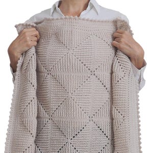 May include: A beige crocheted baby blanket with a square pattern. The blanket is being held up by a person, revealing the intricate details of the crochet work.