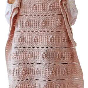 Peut inclure: Une couverture en crochet rose avec un motif floral et des pompons. La couverture a une bordure en franges et est tenue par une personne portant une chemise blanche et un jean bleu. Le texte "DADA'S PLACE" est visible en bas de l'image.
