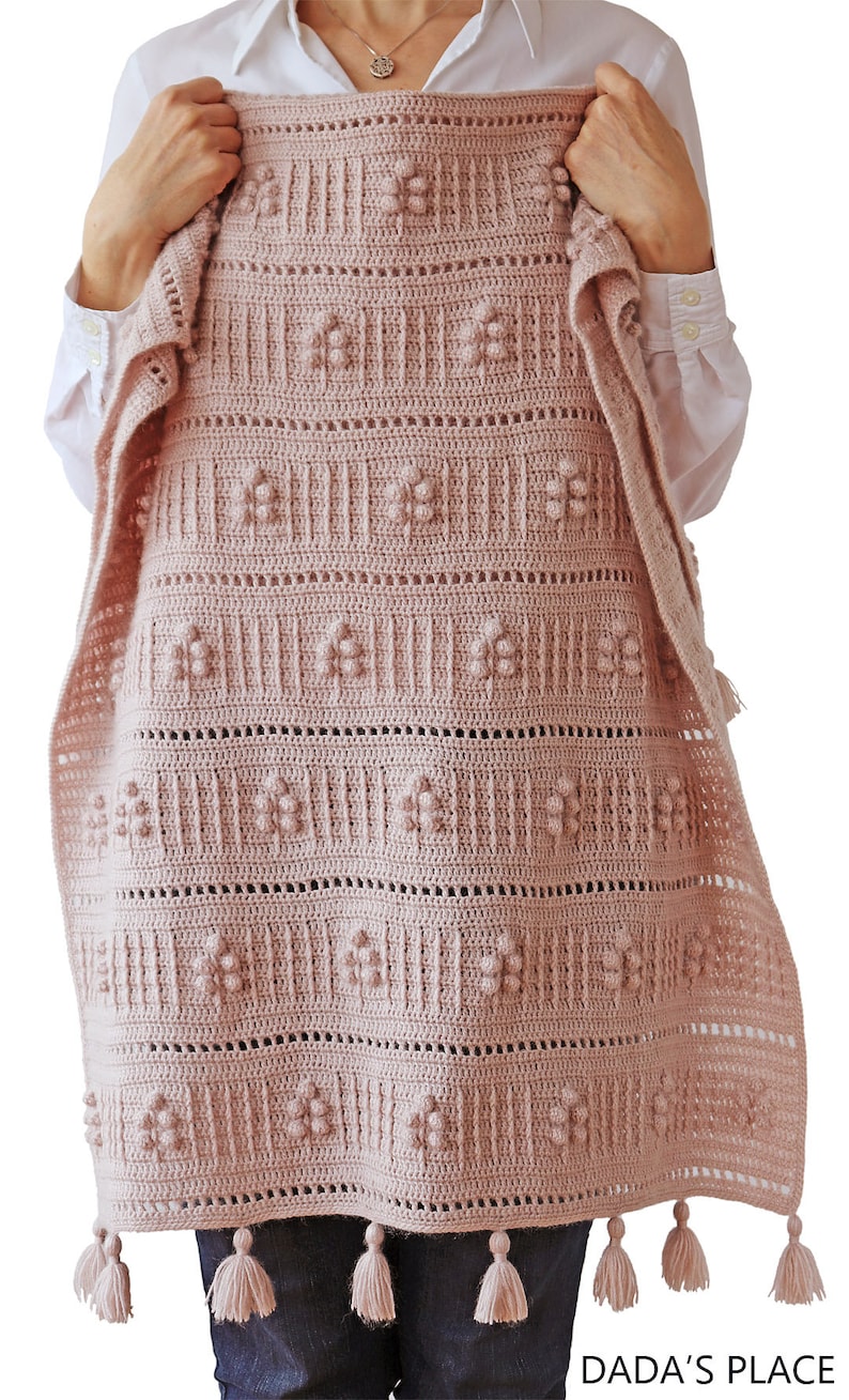 Peut inclure: Une couverture en crochet rose avec un motif floral et des pompons. La couverture a une bordure en franges et est tenue par une personne portant une chemise blanche et un jean bleu. Le texte "DADA'S PLACE" est visible en bas de l'image.