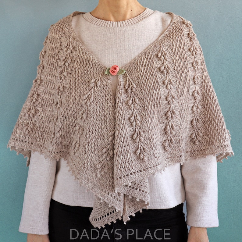 May include: A beige crochet shawl with a delicate leaf pattern and a small pink flower accent. The shawl is draped over a person's shoulders and has a scalloped edge.