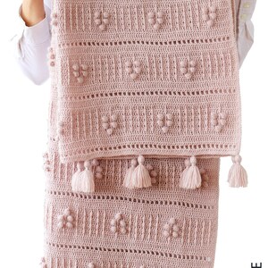 Peut inclure: Une couverture pour b&eacute;b&eacute; en crochet rose avec un motif textur&eacute; et des pompons sur les bords. La couverture est pli&eacute;e en deux et tenue par une personne.