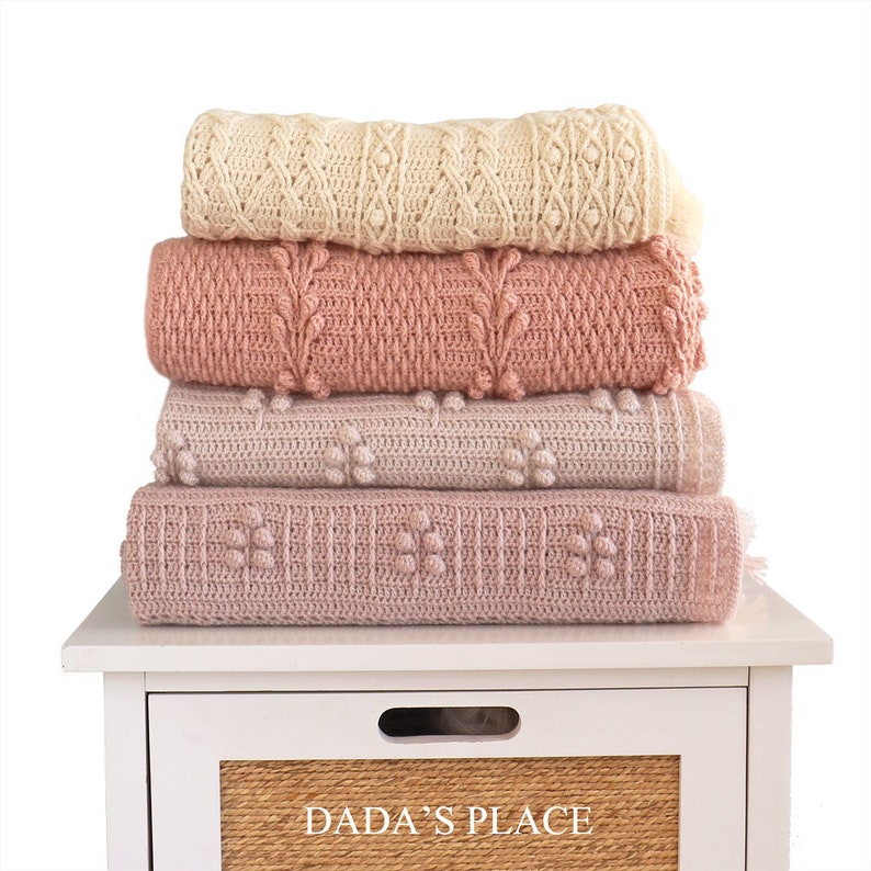 May include: A stack of four crocheted blankets in shades of pink and cream. The blankets are folded and stacked on a white nightstand with a drawer that says "Dada's Place".