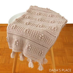 Peut inclure: Une couverture b&eacute;b&eacute; en crochet beige avec un motif textur&eacute; et des franges. La couverture est pli&eacute;e et drap&eacute;e sur un tabouret en bois.