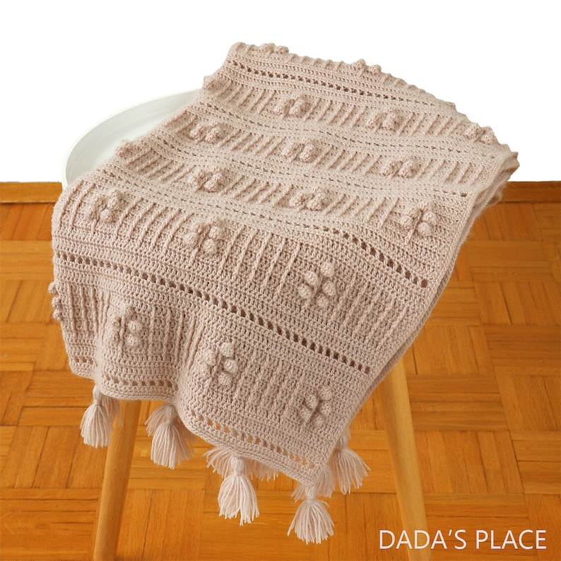 Peut inclure: Une couverture b&eacute;b&eacute; en crochet beige avec un motif textur&eacute; et des franges. La couverture est pli&eacute;e et drap&eacute;e sur un tabouret en bois.