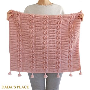 May include: A pink crocheted blanket with a leaf pattern and tassels. The blanket is being held by a person.