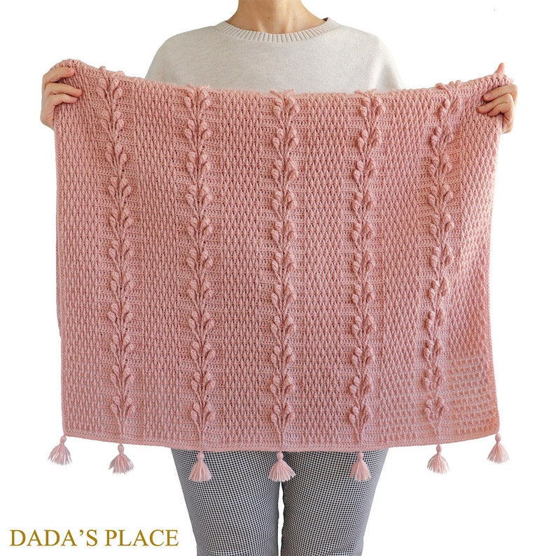 May include: A pink crocheted blanket with a leaf pattern and tassels. The blanket is being held by a person.