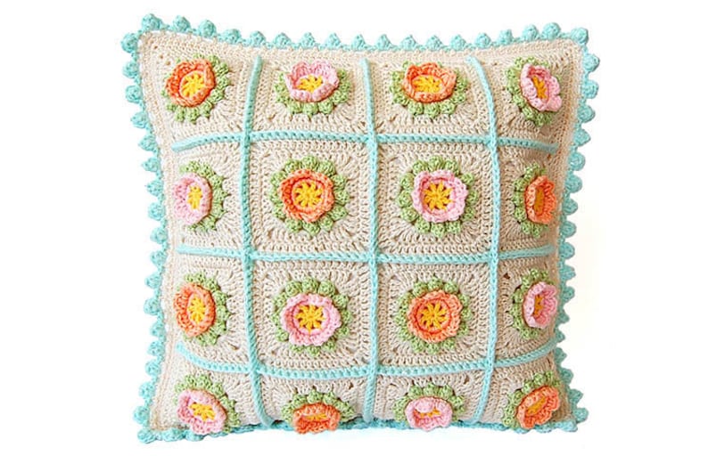 May include: A square crocheted pillow with a floral design. The pillow is made of beige yarn with a light blue border and features a repeating pattern of pink, orange, and yellow flowers.
