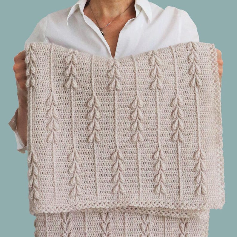 Puede incluir: Una manta rectangular de ganchillo en color beige con un patr&oacute;n vertical repetitivo de racimos en relieve en forma de hoja, separados por columnas rectas de puntos, con un borde texturizado sencillo en los extremos de la tela.