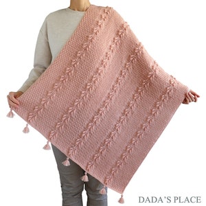 May include: A pink crocheted throw blanket with a leaf pattern and tassels on the edges. The blanket is draped over a person's shoulders.