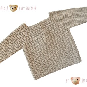May include: A beige baby sweater with a ribbed texture. The sweater has a round neckline and long sleeves. The text "It's a Bear!" and "Baby Sweater" is printed above the sweater. The text "By Dada's Place" is printed below the sweater.