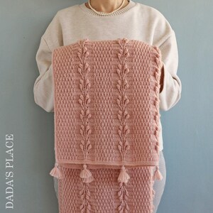 May include: A pink crocheted baby blanket with a leaf pattern and tassels.