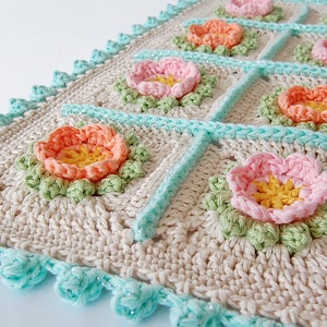 May include: A crocheted blanket with a floral design. The blanket is made with beige yarn and features a repeating pattern of pink, orange, and yellow flowers. The edges of the blanket are trimmed with a turquoise crocheted border.